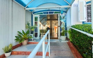 L’entrée d’un hôtel, avec un passage couvert, des plantes en pot et un comptoir d’accueil.