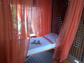 Ein Einzelbett mit weißer Matratze und orangefarbenen Akzenten, umrahmt von einem Holzgestell und einem roten Moskitonetz.