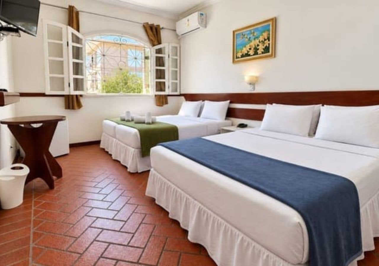 A hotel room with a bed, a small table, a chair, a window with curtains, and a painting on the wall.