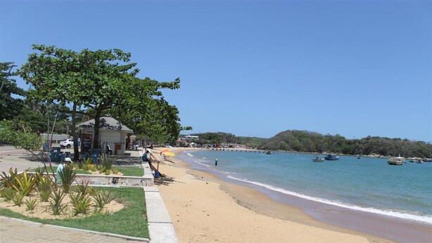 A beach with a paved walkway, a building with a sign, and boats in the water. - Pousada da Lenna (Rio das Ostras)