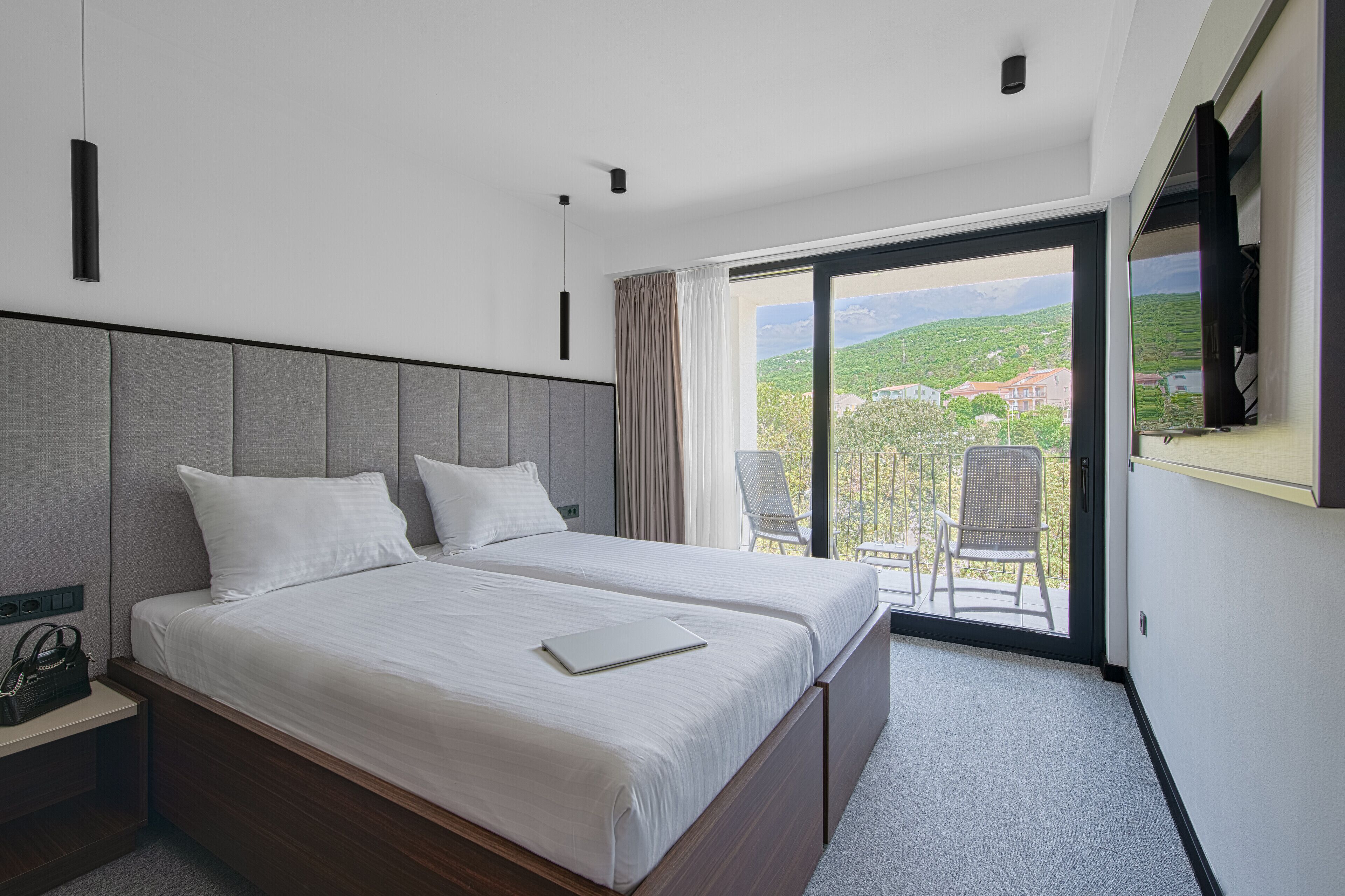 A double bed with a laptop, a nightstand, a chair, and a balcony with a view of a green landscape.