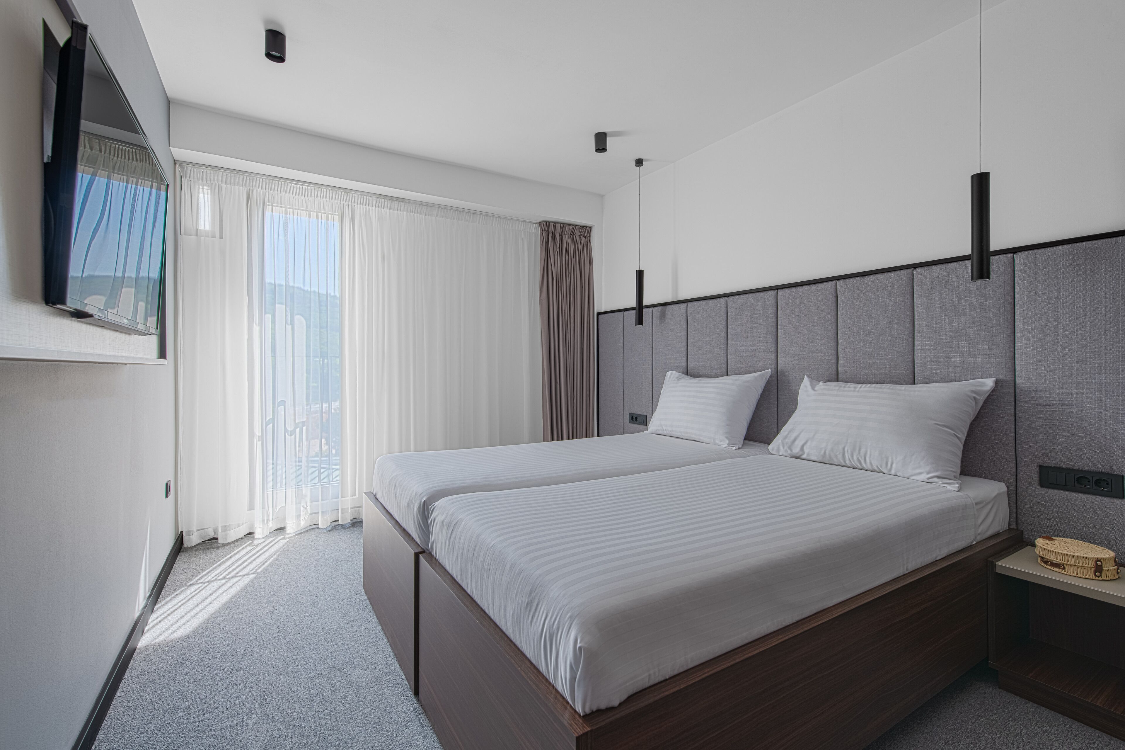 A modern hotel room with a large bed, a TV mounted on the wall, and a view of the city through the window.