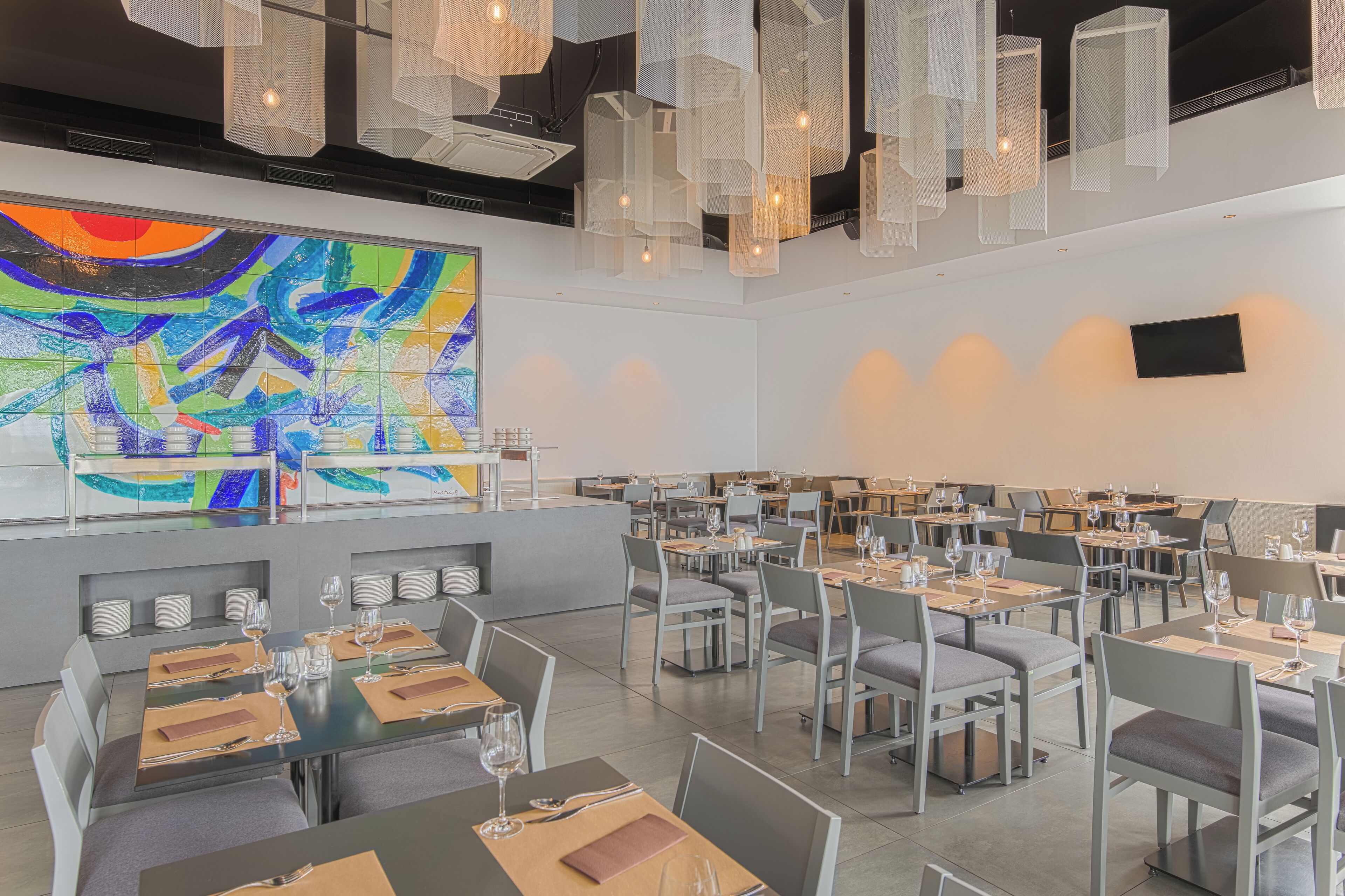 A modern dining area with tables set for meals, a large abstract painting on the wall, and hanging pendant lights.