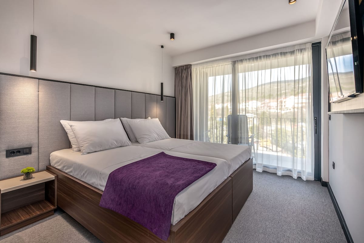 A modern bedroom with a large bed, a nightstand, a TV, and a view of the outdoors through the window.