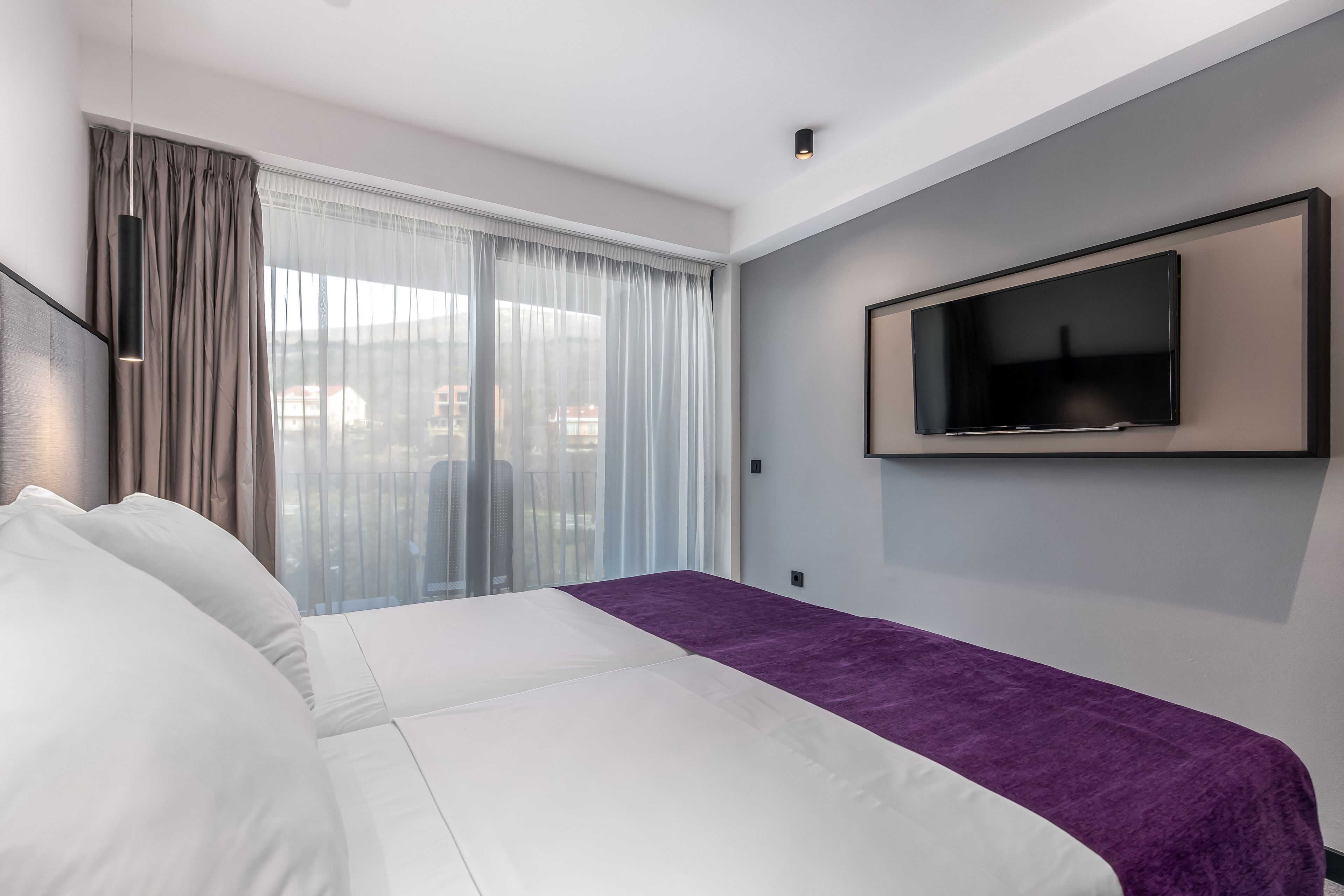 A modern hotel room with a large bed, a flat-screen TV mounted on the wall, and a view of the city through sheer curtains.