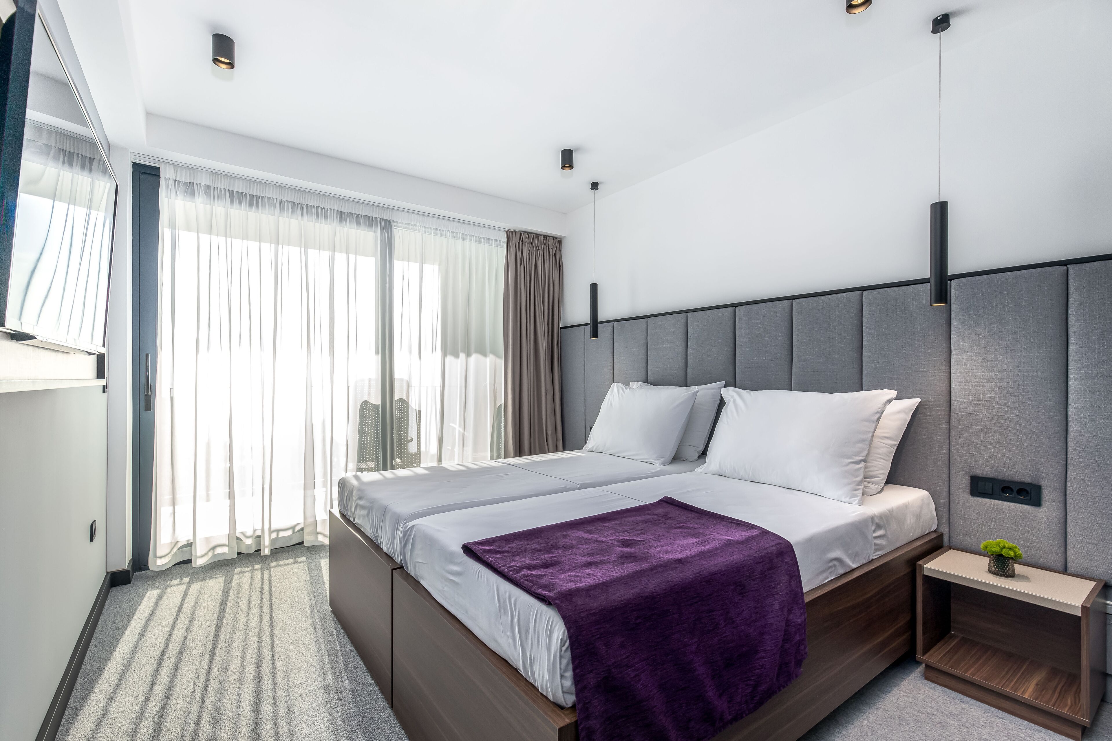 Una habitación de hotel moderna con una cama grande, una mesita de noche con una planta y vistas a la ciudad a través de cortinas translúcidas.