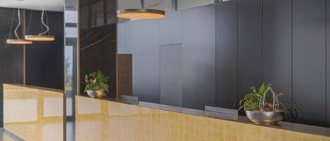 Zona de recepción con mostrador de madera, una planta en maceta e iluminación colgante.