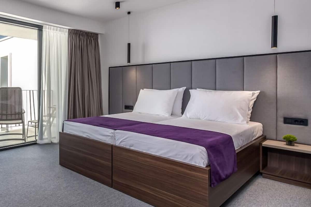 A modern hotel room with a large bed, a wooden headboard, a small bedside table, and a view of the balcony.