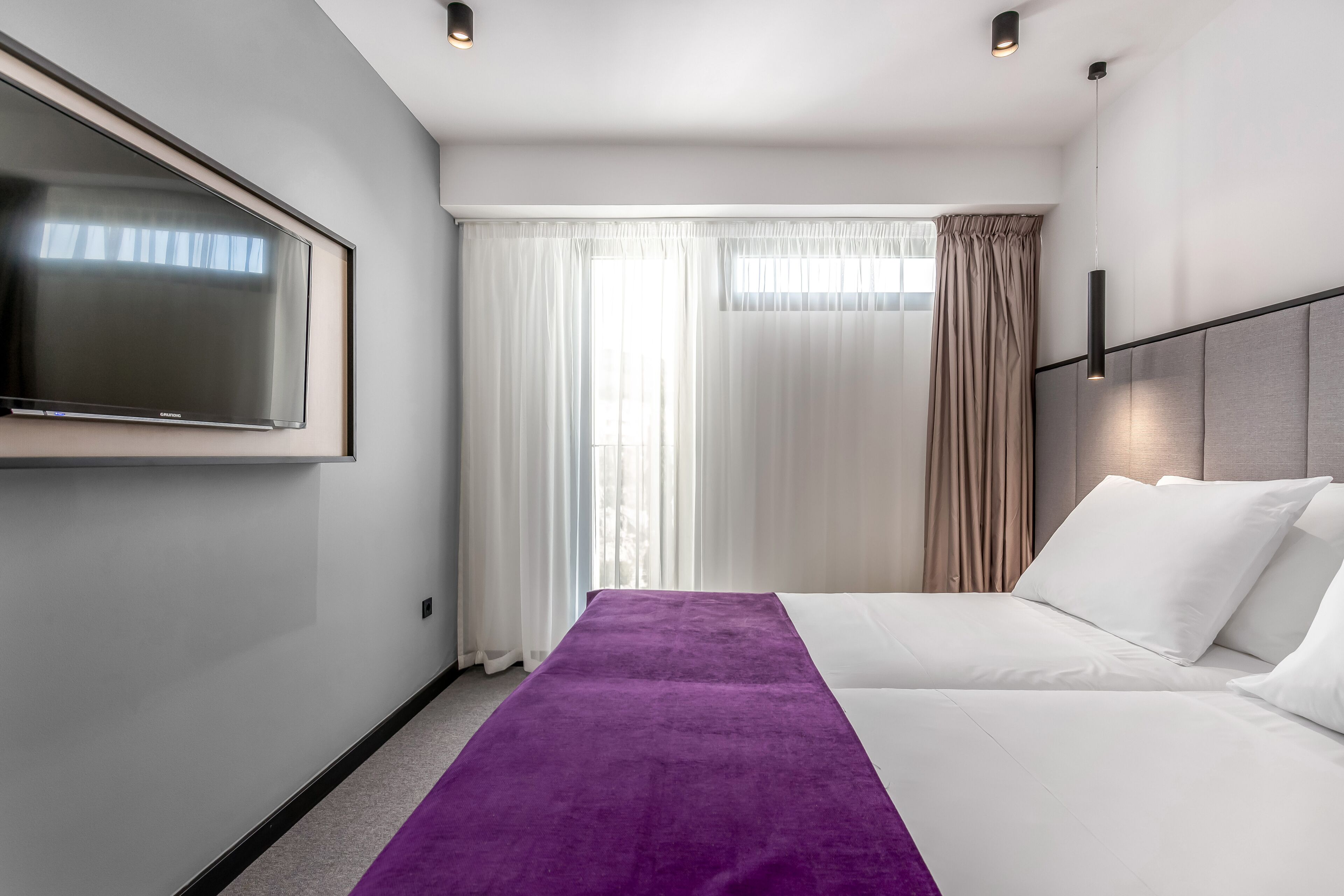 Habitación de hotel con una televisión de pantalla plana grande, una cama con ropa blanca, un plaid morado y una ventana con cortinas translúcidas.