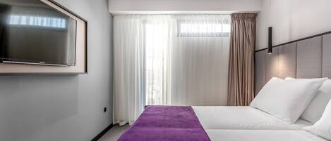 Habitación de hotel con una televisión de pantalla plana grande, una cama con ropa blanca, un plaid morado y una ventana con cortinas translúcidas.