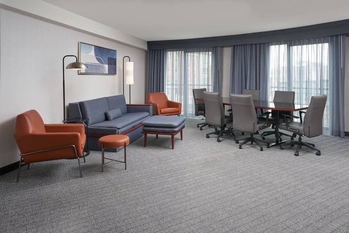 A modern hotel room with a gray carpet, a gray sofa, orange armchairs, a wooden coffee table, and a conference table with chairs.