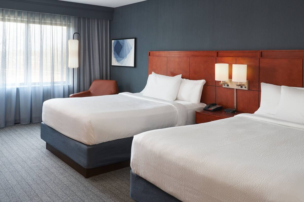 A hotel room with two beds, a chair, a nightstand with a phone, and a wall-mounted lamp.