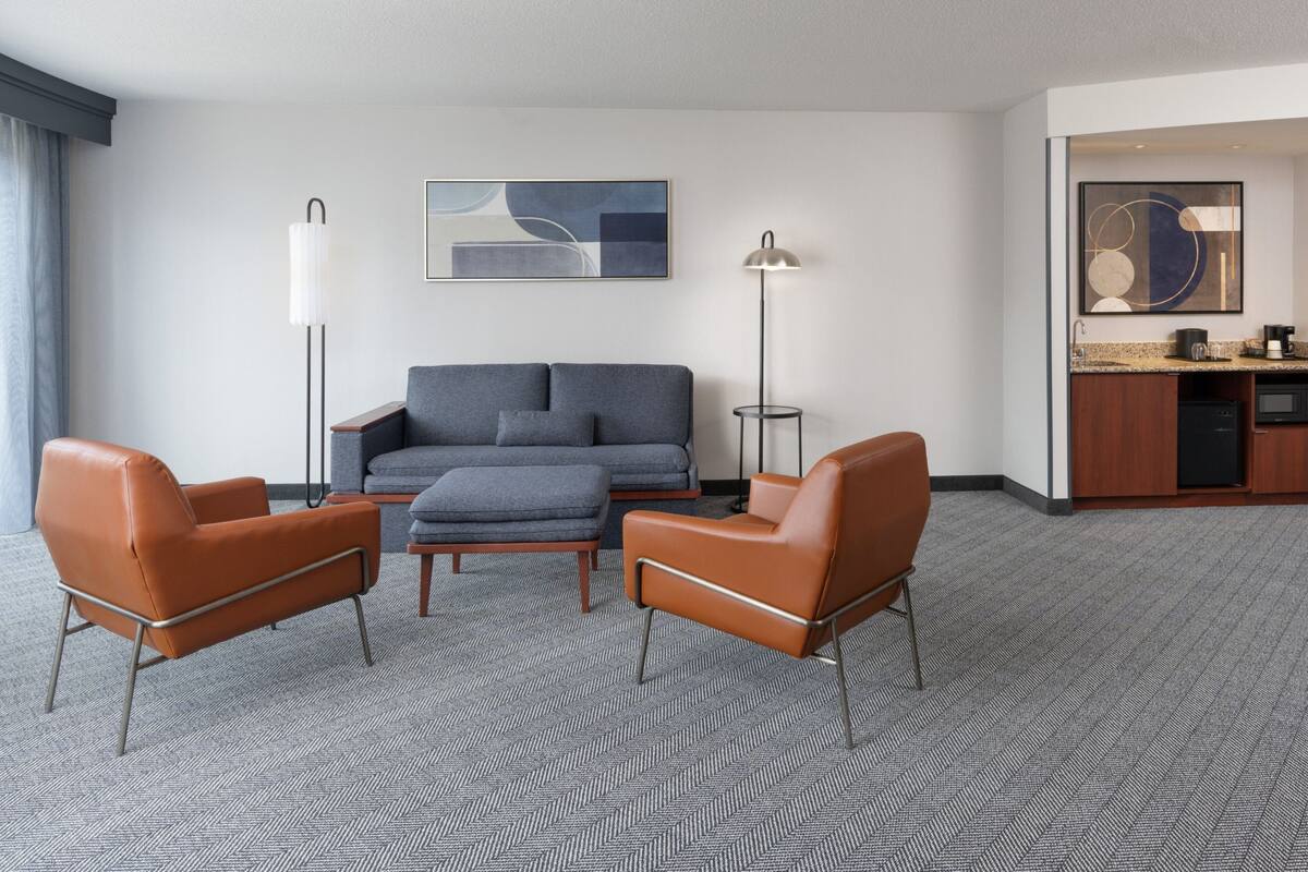 A modern hotel room with a grey sofa, two leather armchairs, a coffee table, a floor lamp, and a painting on the wall.