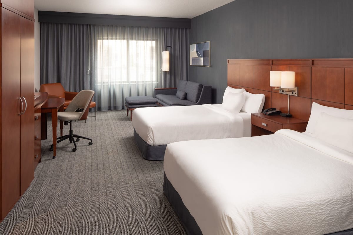 A hotel room with two beds, a desk, a chair, a sofa, and a lamp.