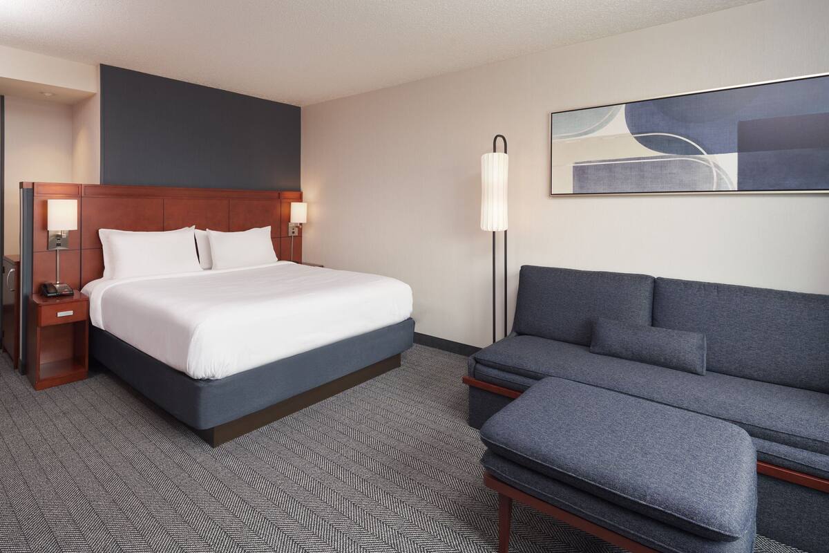A hotel room with a bed, a sofa, a nightstand, a lamp, and a painting on the wall.