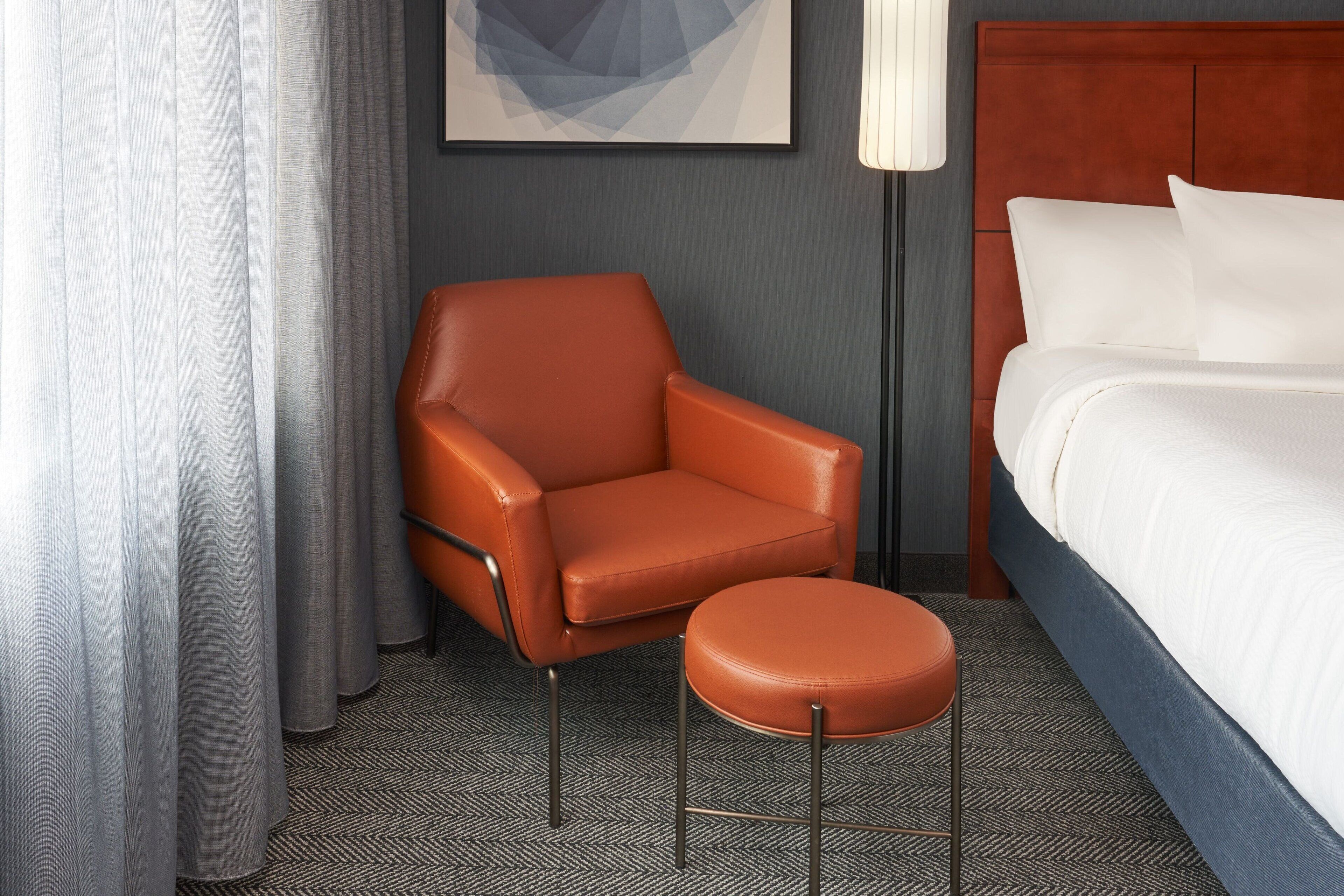 A hotel room with a brown leather chair, a matching ottoman, a bed with white linens, a grey wall, and a framed abstract artwork above the bed.