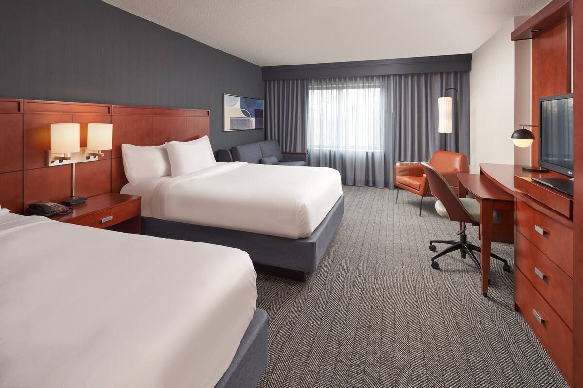 A hotel room with two beds, a desk, a chair, a television, and a large window with curtains.