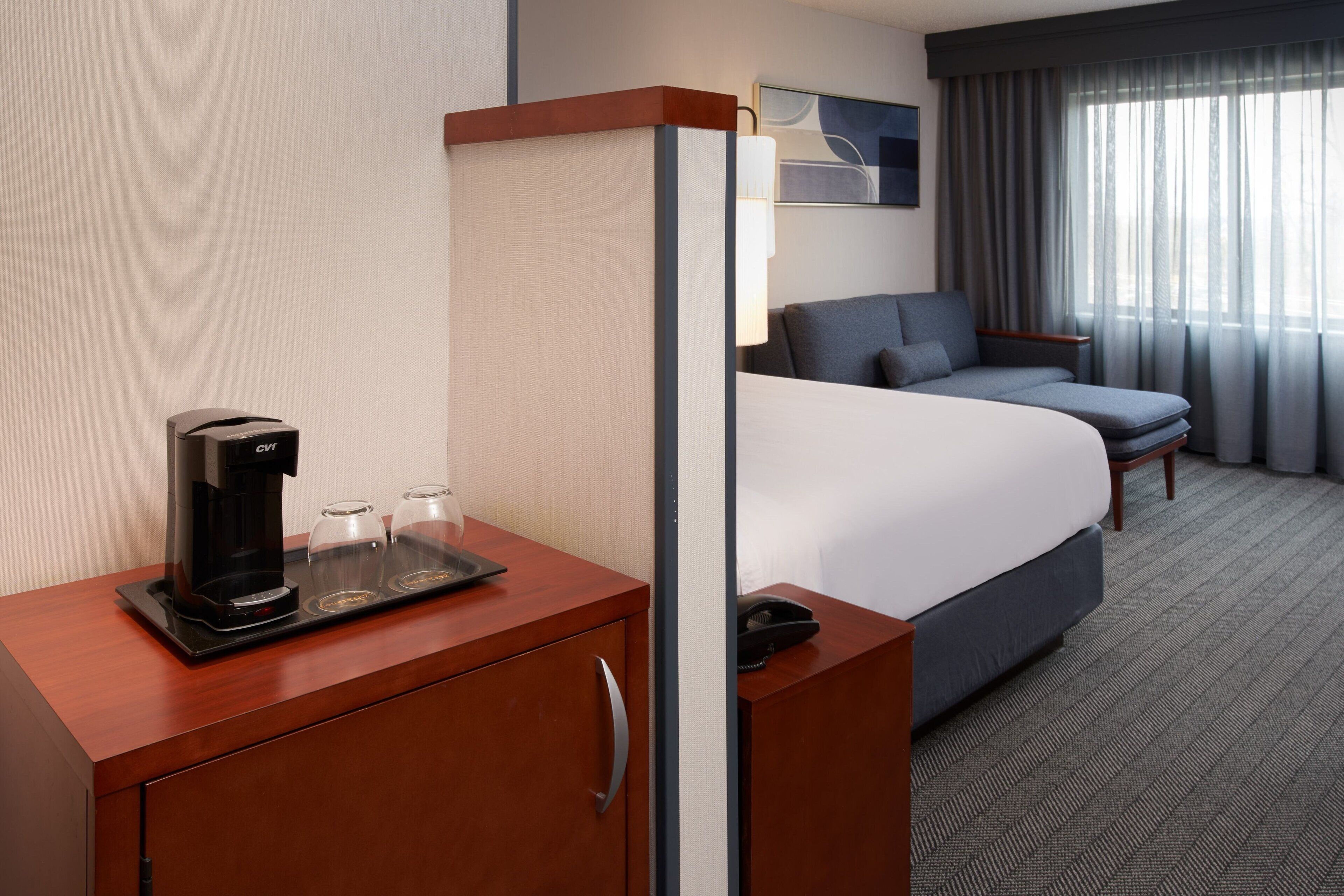A hotel room with a bed, a desk with a coffee maker and glasses, a phone, and a view of the outside through a window.