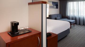 A hotel room with a bed, a desk with a coffee maker and glasses, a phone, and a view of the outside through a window.