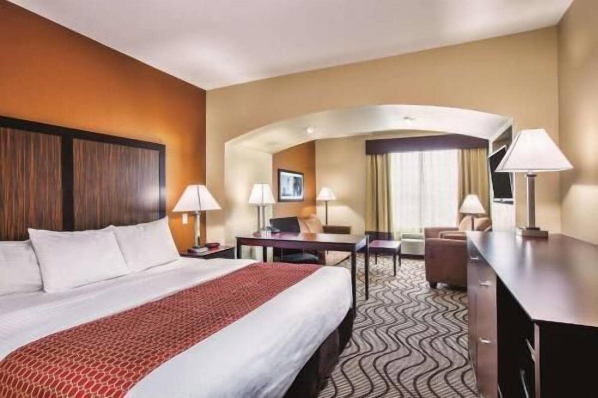 A hotel room with a bed, desk, chair, lamps, and a view of the hallway.