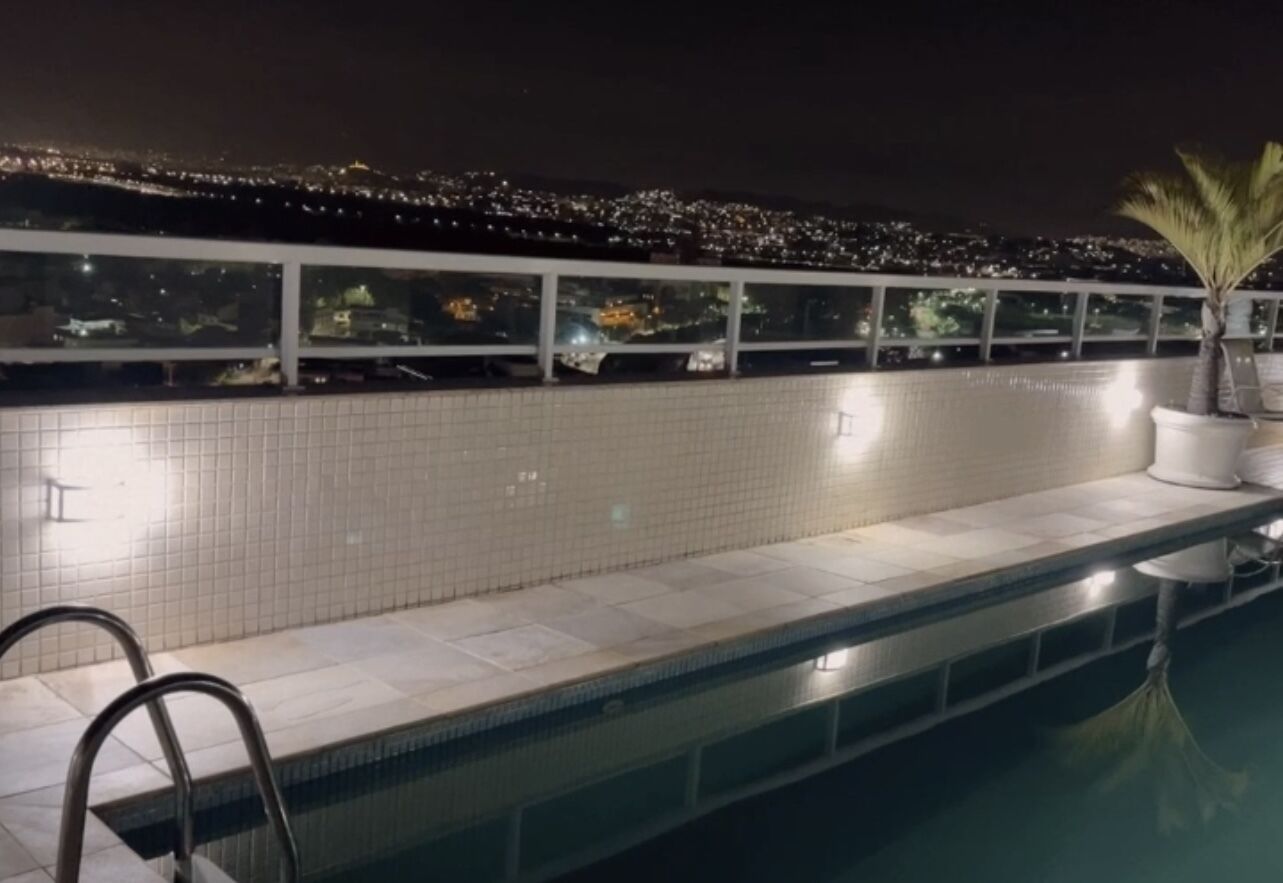 Une piscine avec un bord carrelé et un escalier en métal. Une rambarde offre une vue sur le paysage urbain la nuit.