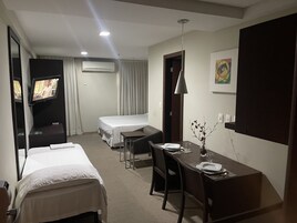 A hotel room with a bed, a desk with a chair, a TV, an air conditioning unit, and a painting on the wall.