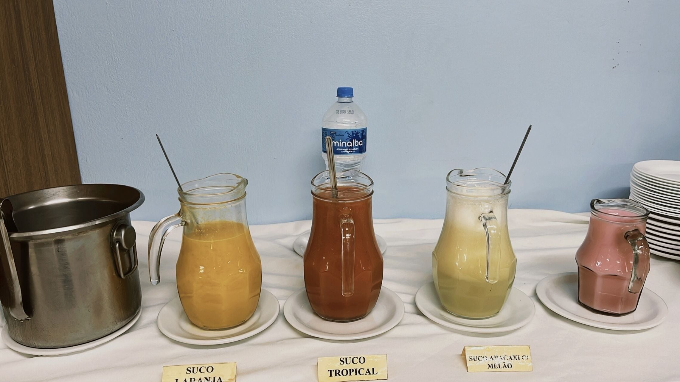 Una estación de bebidas con varias opciones, incluyendo zumos como Suco Jarania, Suco Tropical y Suco Apacaici Melão.