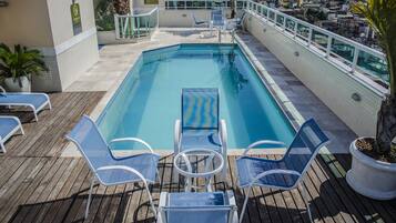 A rooftop pool with blue water, surrounded by white lounge chairs and a small table.