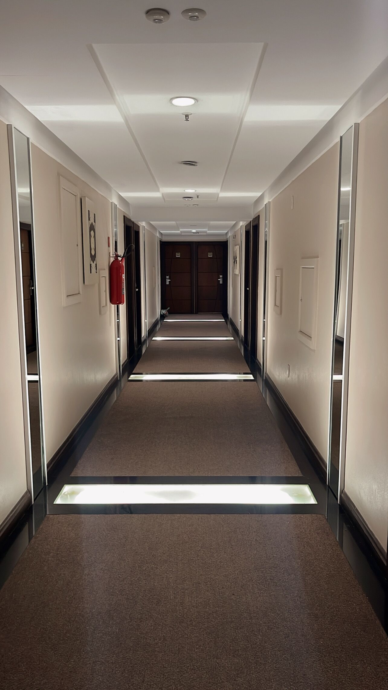 A long, narrow hotel corridor with a carpeted floor, recessed ceiling lights, and doors on both sides.