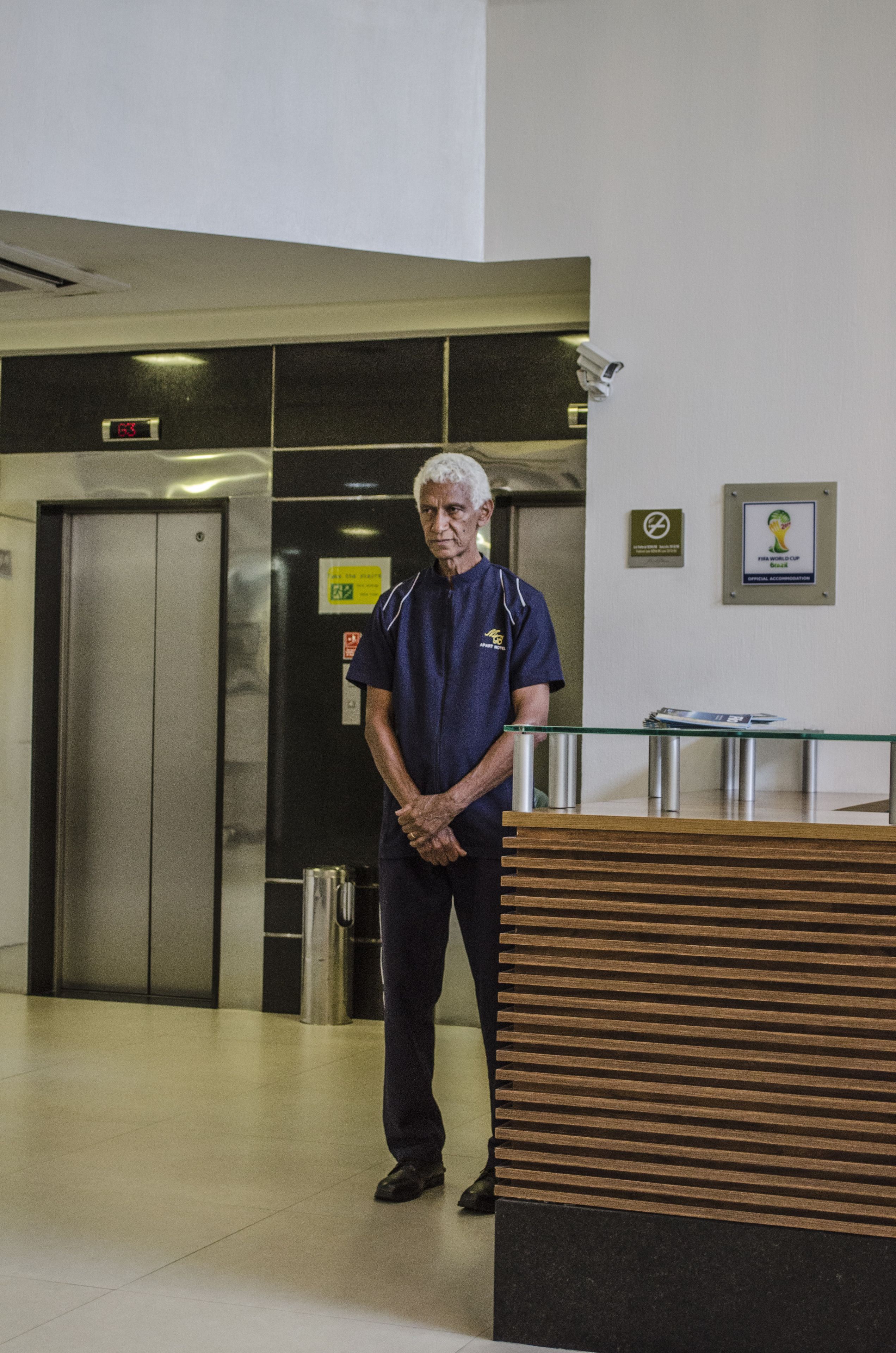 Un hombre de pie en un vestíbulo con un ascensor, un mostrador de recepción de cristal y el logotipo enmarcado de la Copa Mundial de la FIFA en la pared.