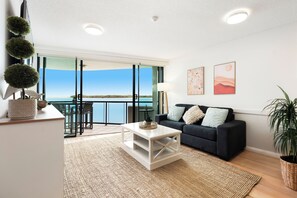 A modern living room with a balcony, a white coffee table, a dark sofa, and a large window offering a view of the sea.