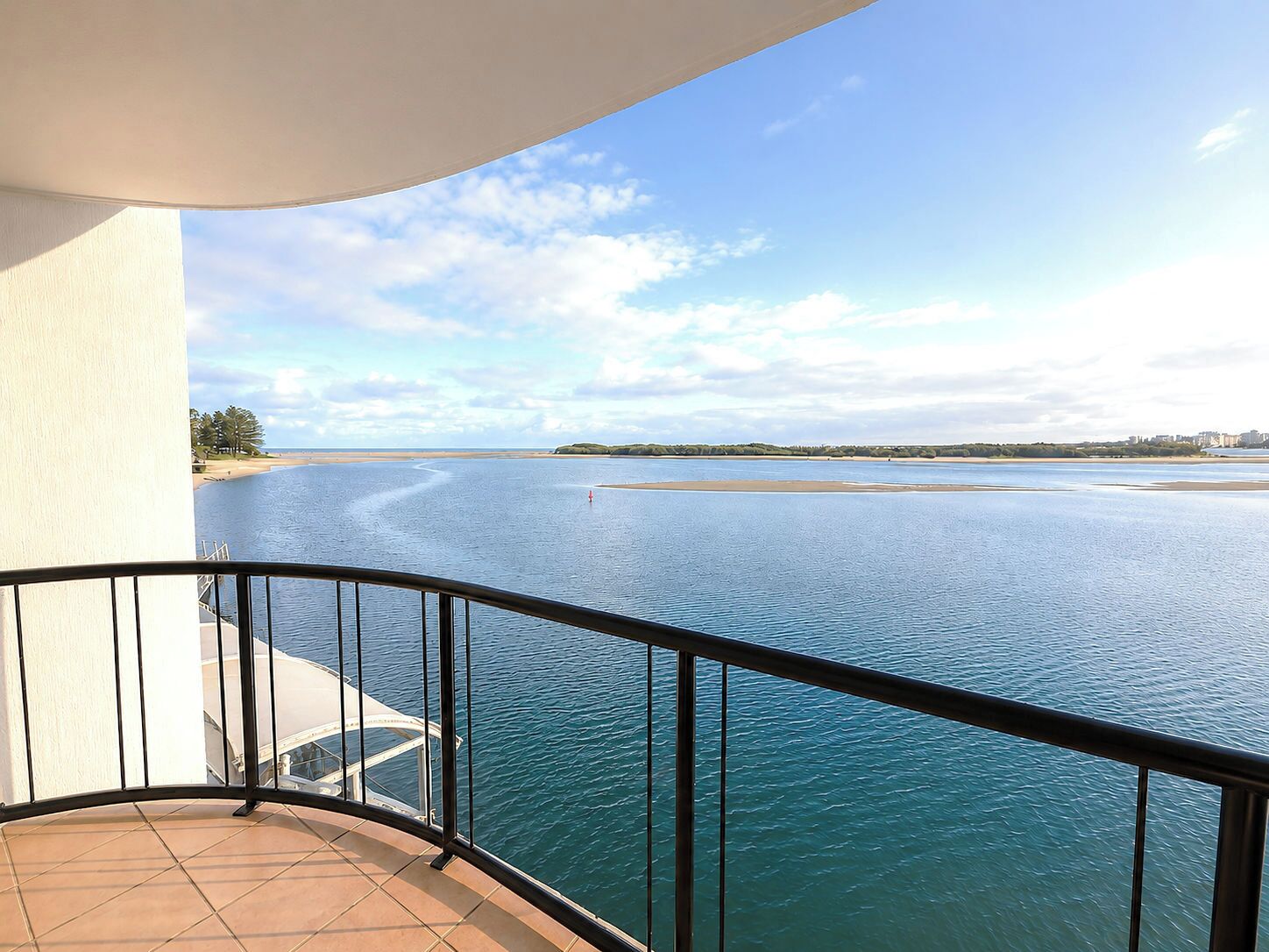 A balcony with a view of a calm body of water, a sandy beach, and a distant shoreline with buildings.
