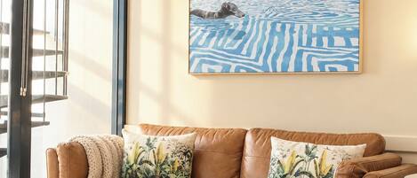 A living room with a brown leather sofa, two round wooden side tables, a patterned rug, and a large abstract painting on the wall.