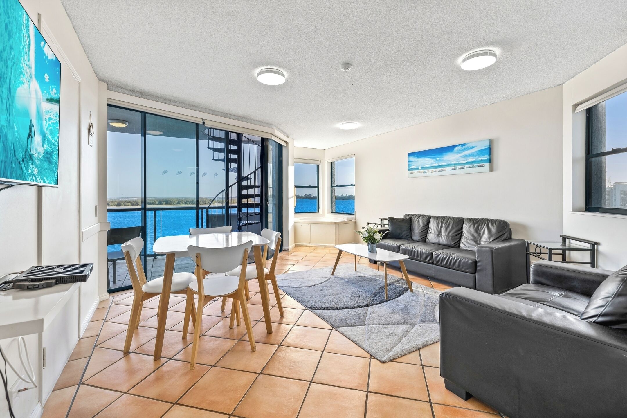 A spacious living area with a large window offering a view of the sea, a black leather sofa, a white dining table with chairs, and a small coffee table.