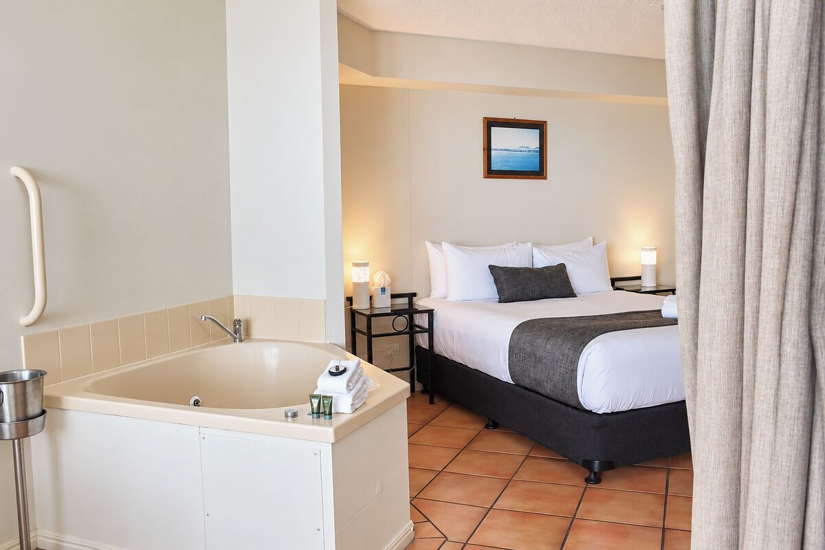 A hotel room with a bathtub, a bed with white bedding, a nightstand with a lamp, and a picture on the wall.