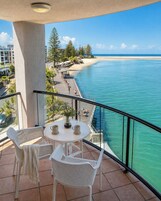 A balcony with a view of a beach and ocean, featuring a table with chairs and a vase.