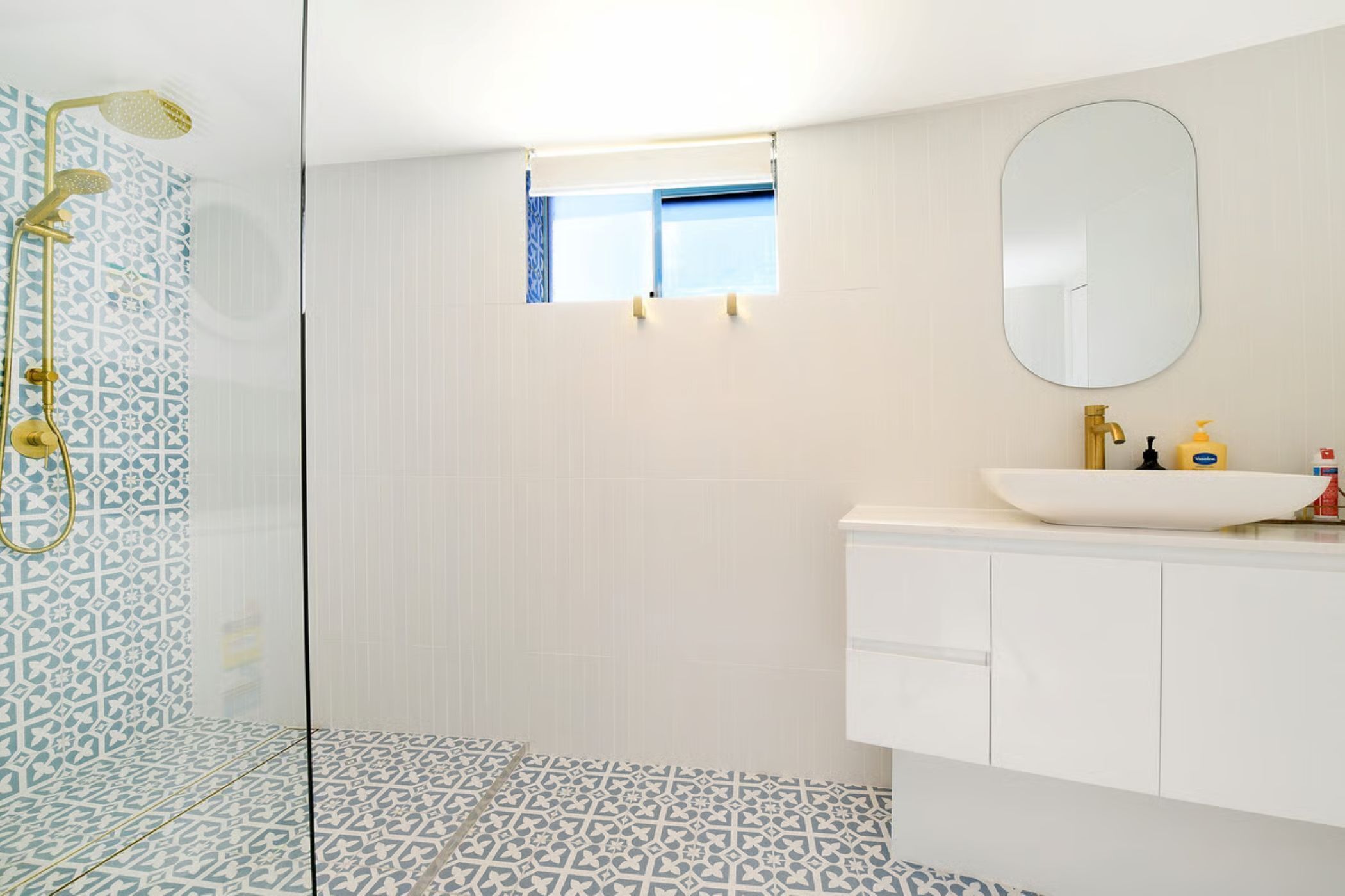 A bathroom with patterned tiles, a glass-enclosed shower, a white sink with gold fixtures, and a small window above the mirror.