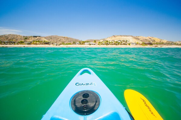 Un kayak con el nombre de marca 'Gemini' en la proa, dirigiéndose hacia una playa con aguas turquesas cristalinas y colinas y edificios al fondo.