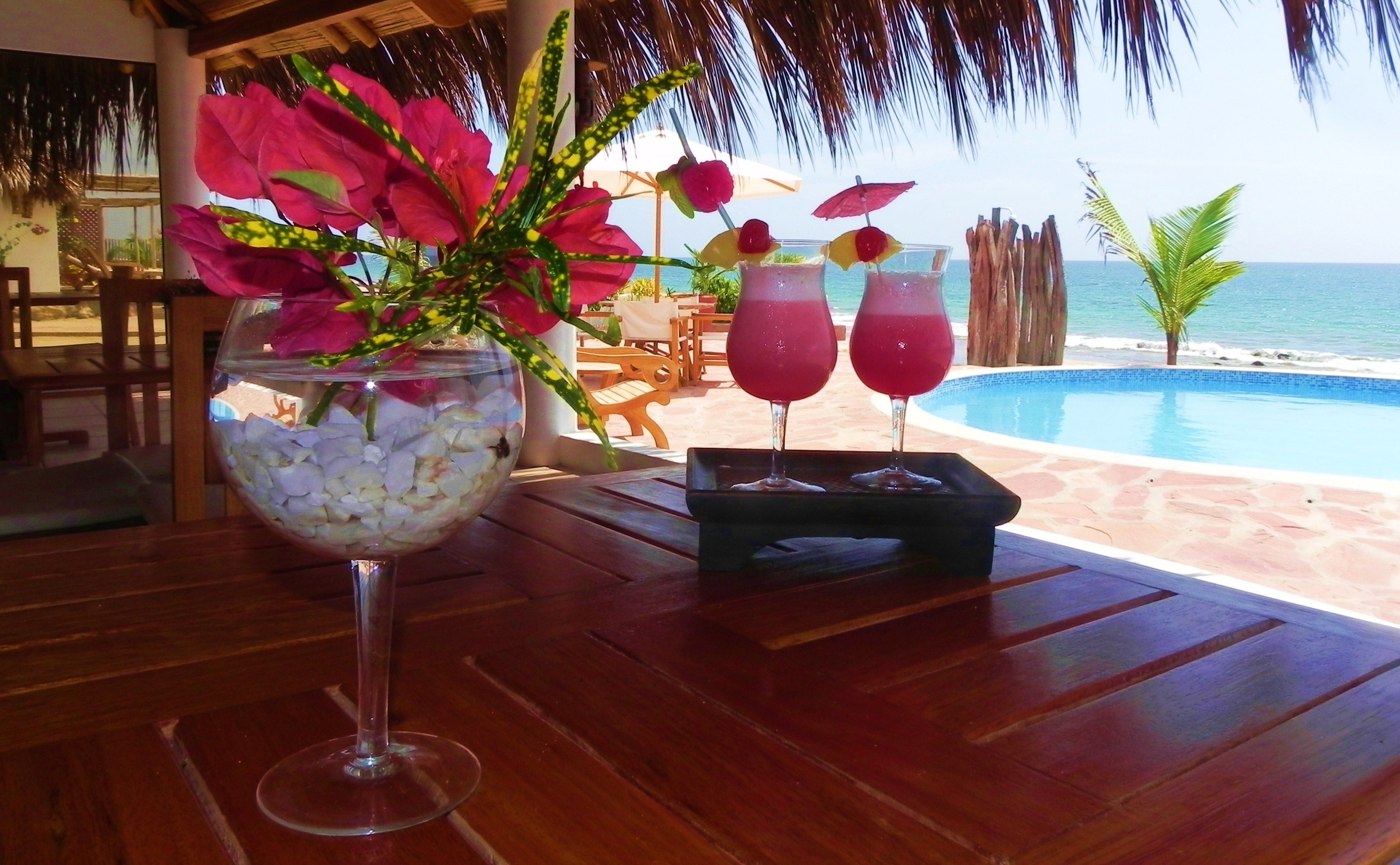 A table with a glass of drink, two cocktails, a flower arrangement, and a view of the ocean.
