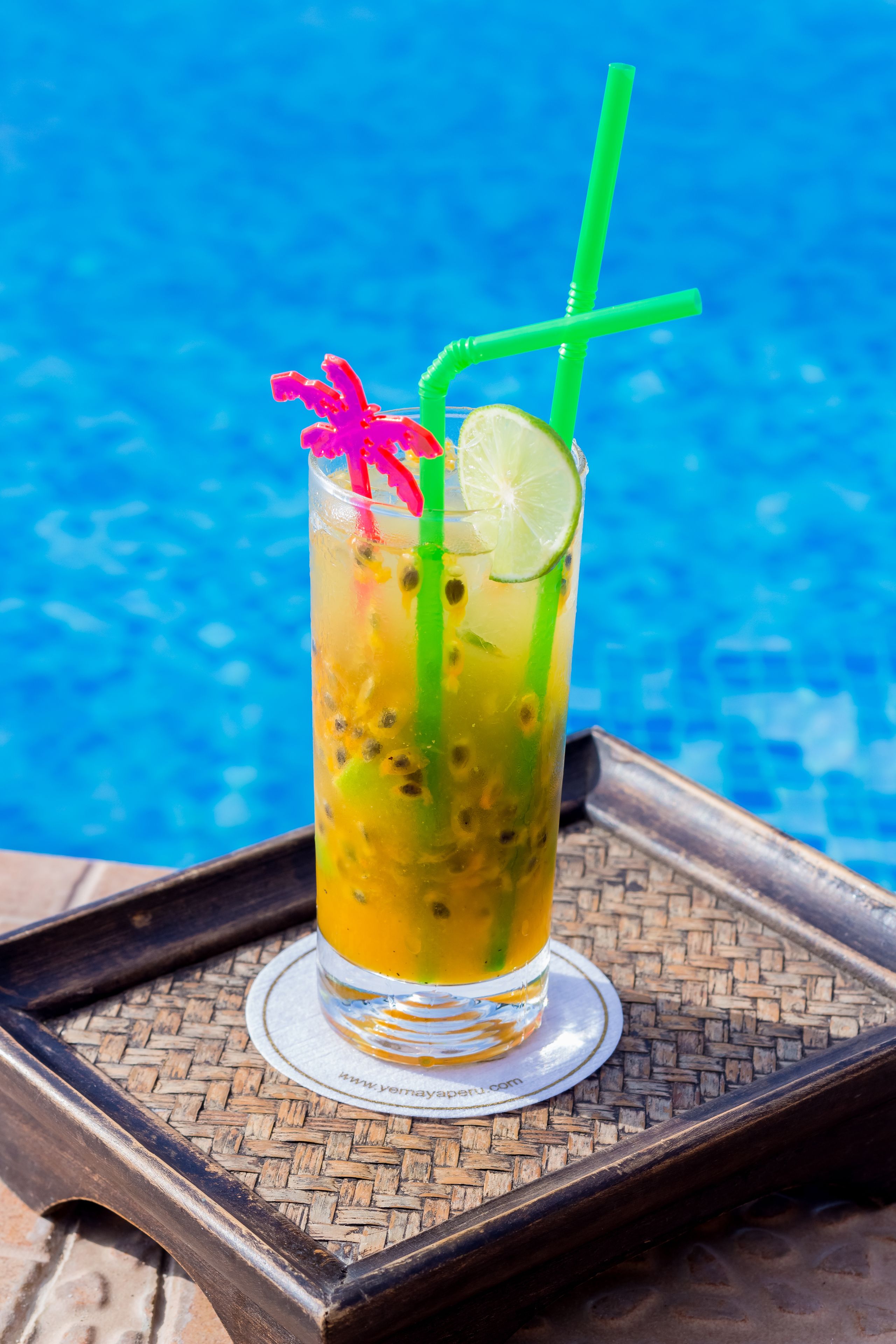 A glass of tropical drink with a lime slice and a green straw, placed on a woven tray by a pool.