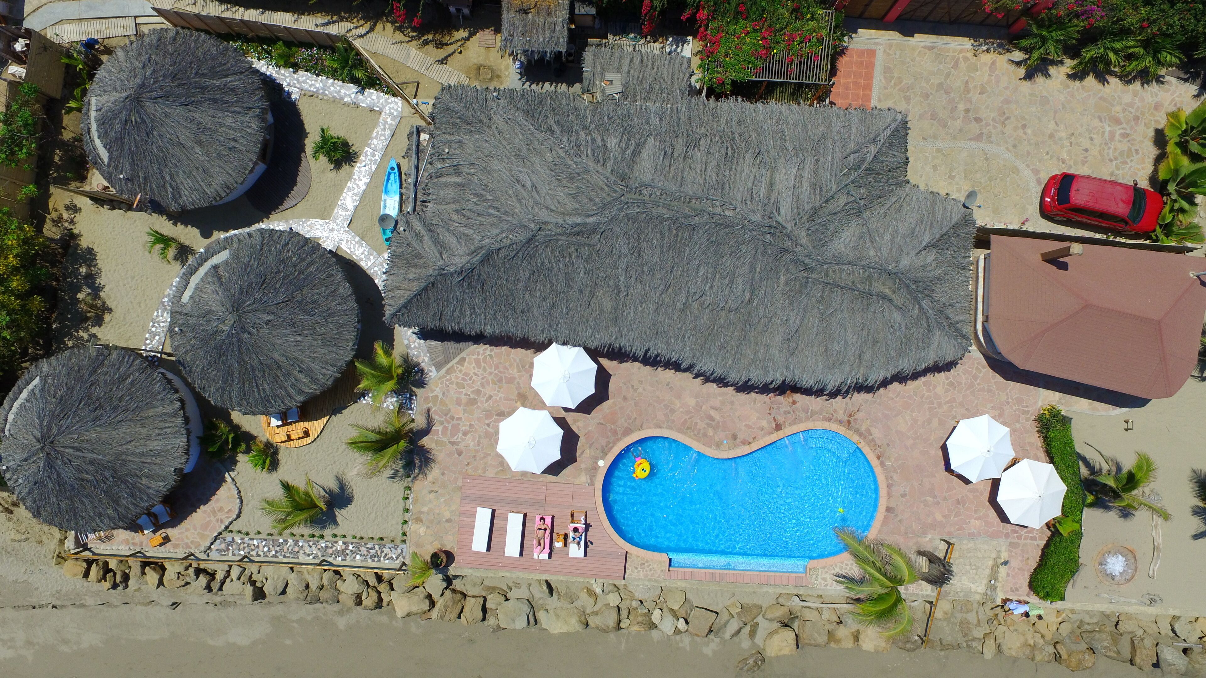 Vue aérienne d’un complexe hôtelier avec une piscine, des parasols en chaume et une voiture rouge garée à proximité.