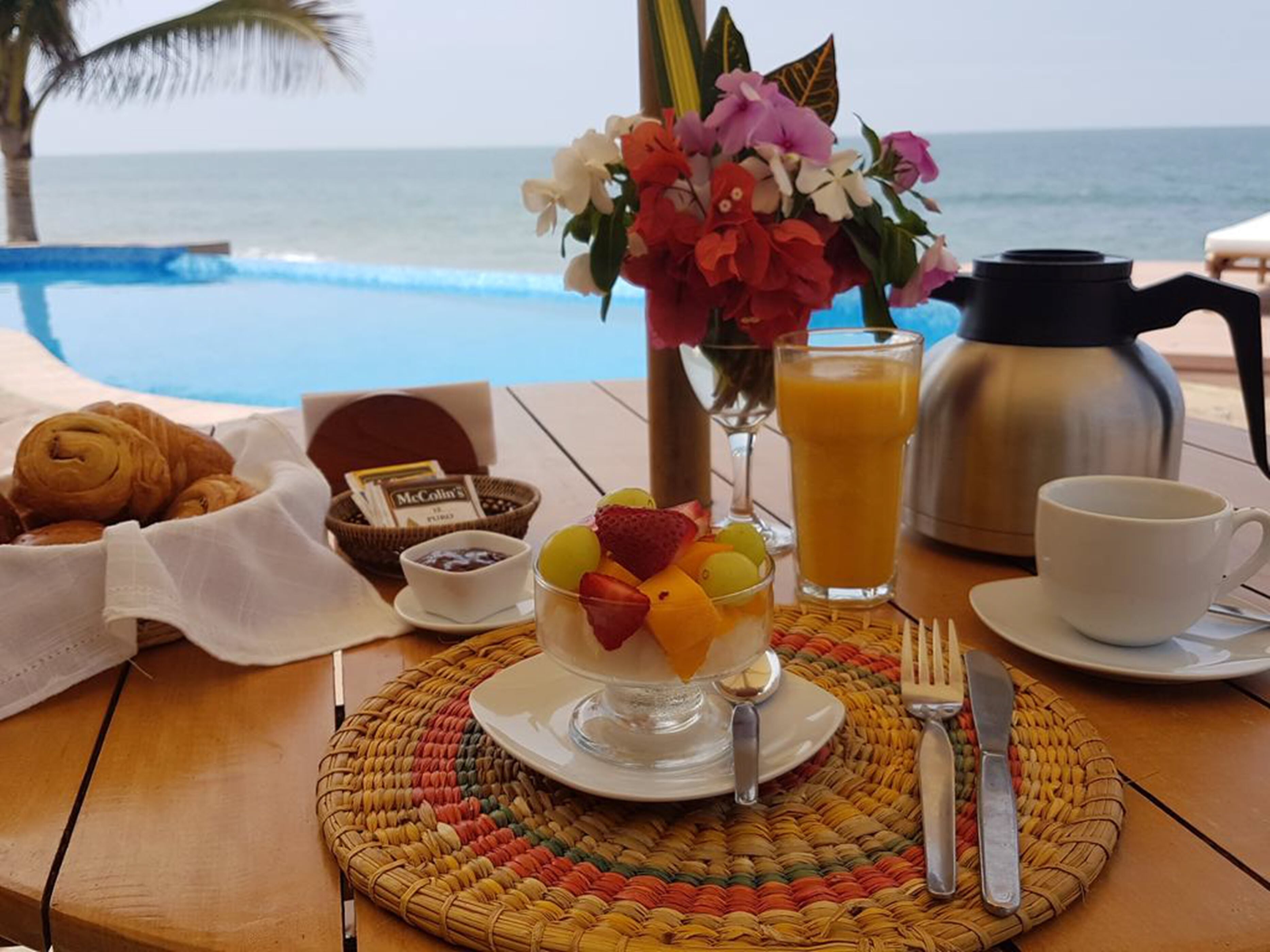 A breakfast setup with croissants, a fruit bowl, a coffee pot, and a view of the ocean.