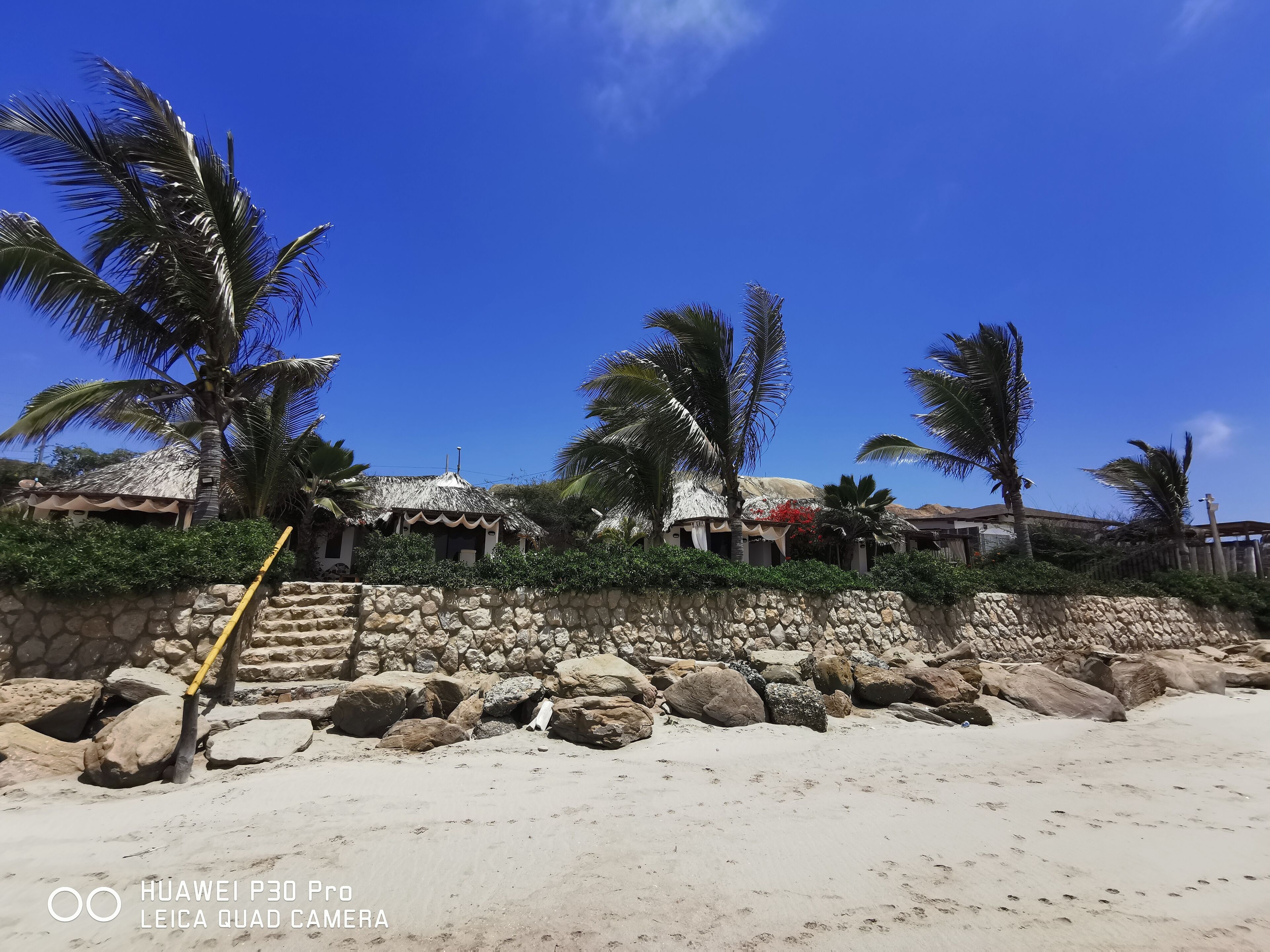 Una propiedad frente a la playa con muros de piedra, palmeras y edificios con techo de paja.
