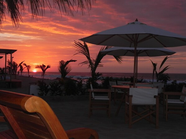 A beachside patio with wooden chairs and umbrellas, overlooking the ocean at sunset.