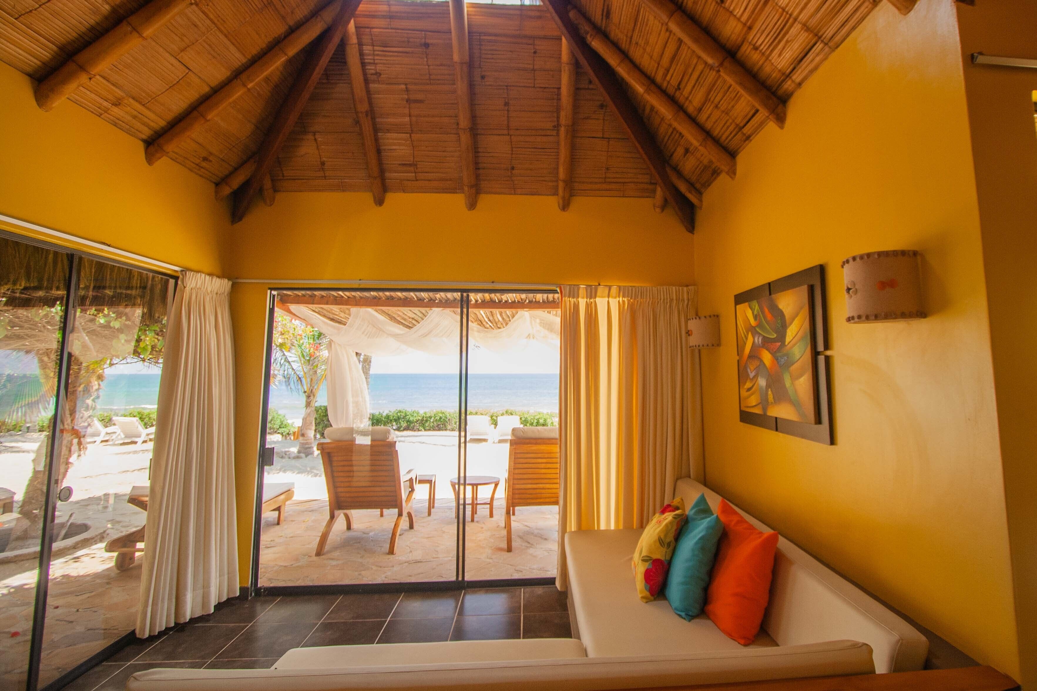 A room with a view of the beach, a sofa with colorful cushions, and a painting on the wall.