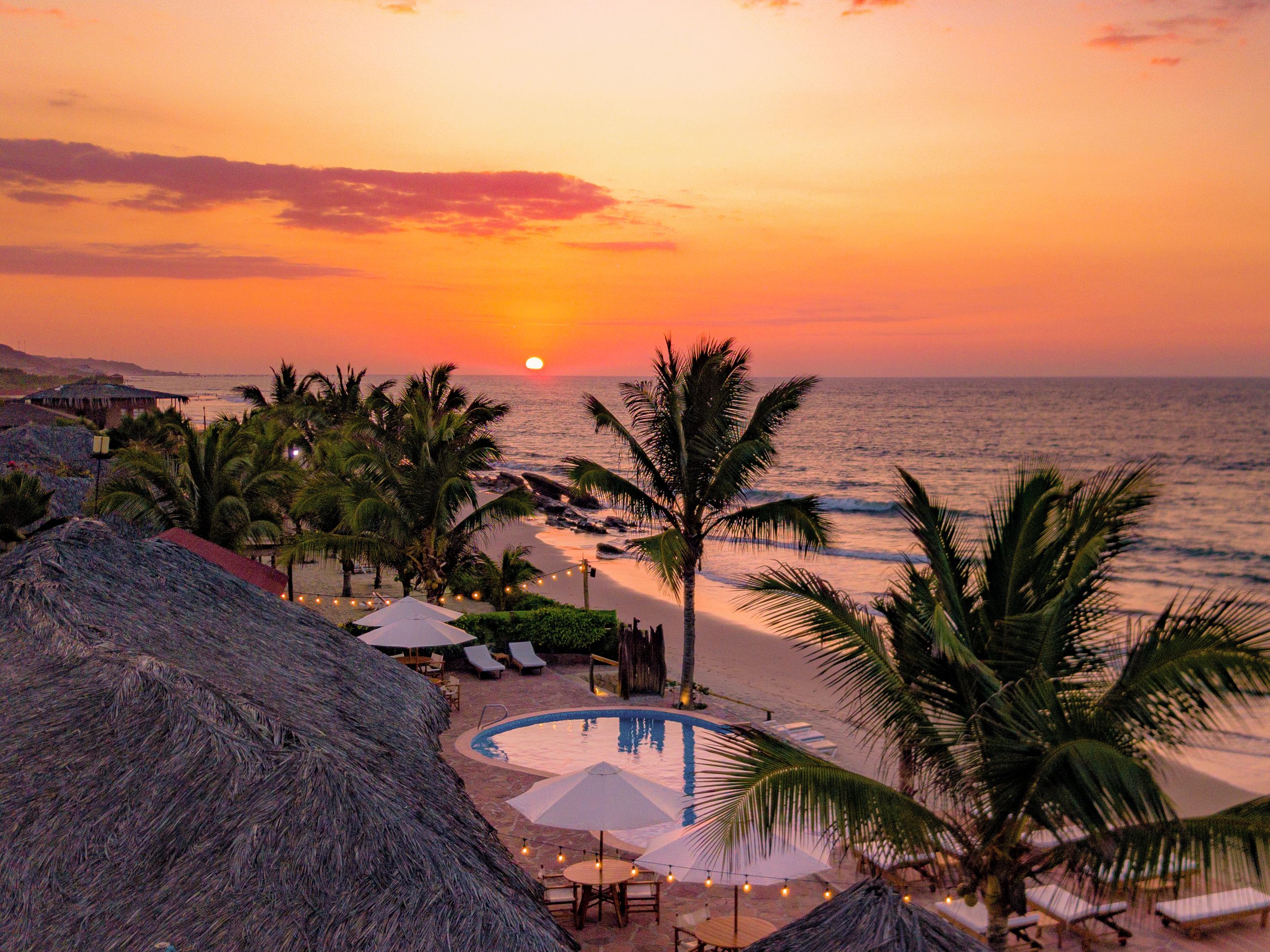 A resort with a pool, beach chairs, umbrellas, and palm trees.