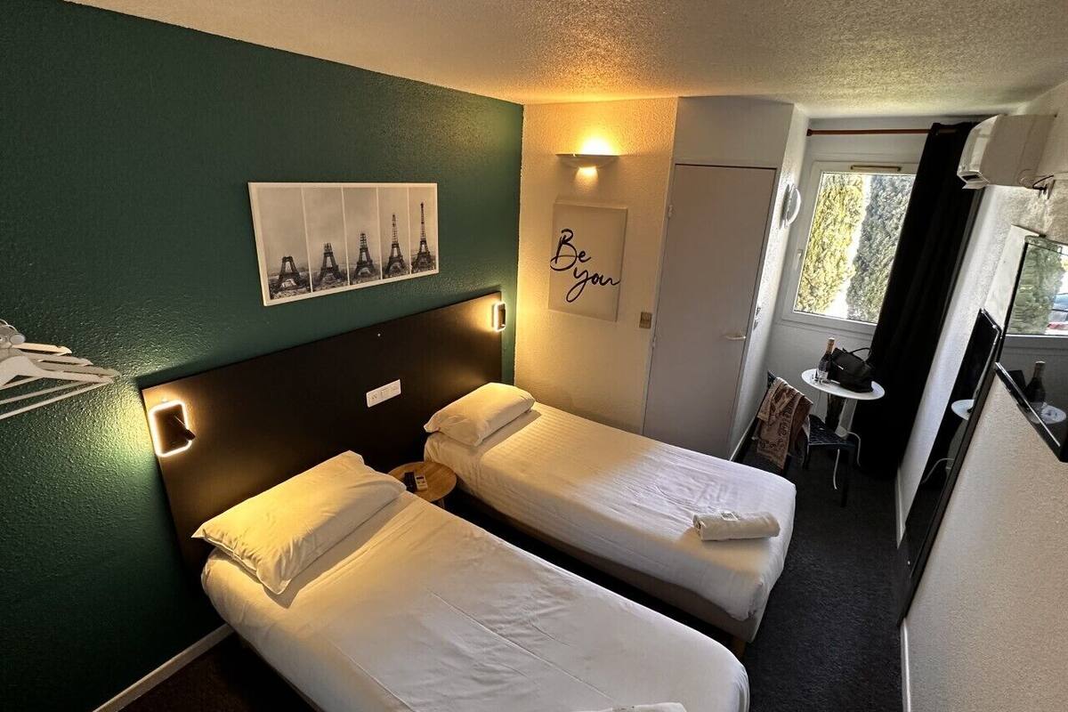 A hotel room with two single beds, a small desk, a chair, and a window with a view of trees.