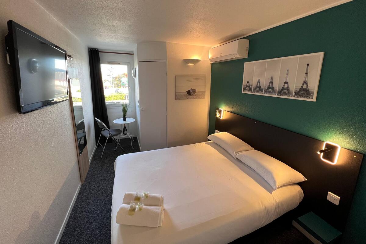 A hotel room with a bed, a TV, a small table with a chair, and a window.