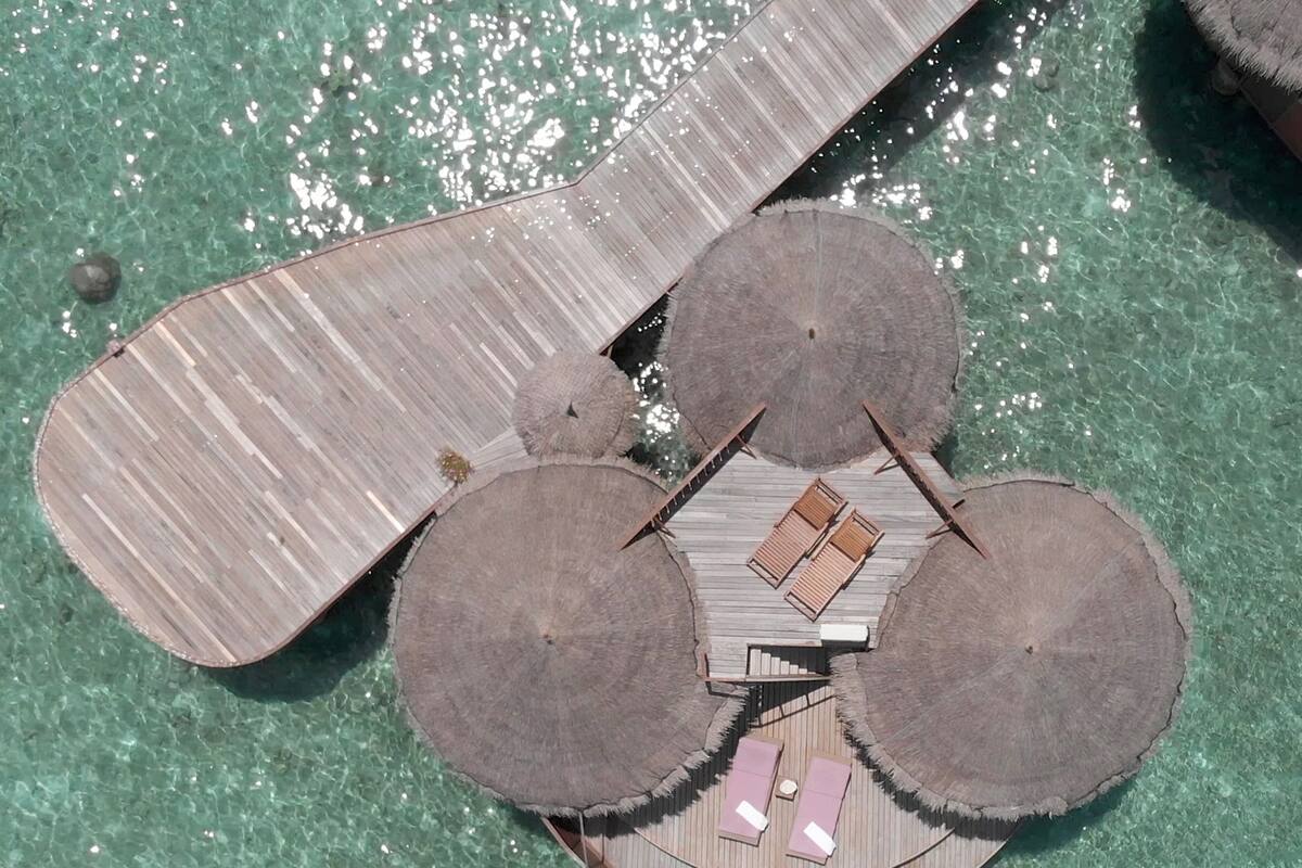 Vue aérienne d’un complexe hôtelier doté d’une structure circulaire centrale, entourée d’espaces de détente et d’une grande terrasse en bois qui s’étend au-dessus de l’eau.