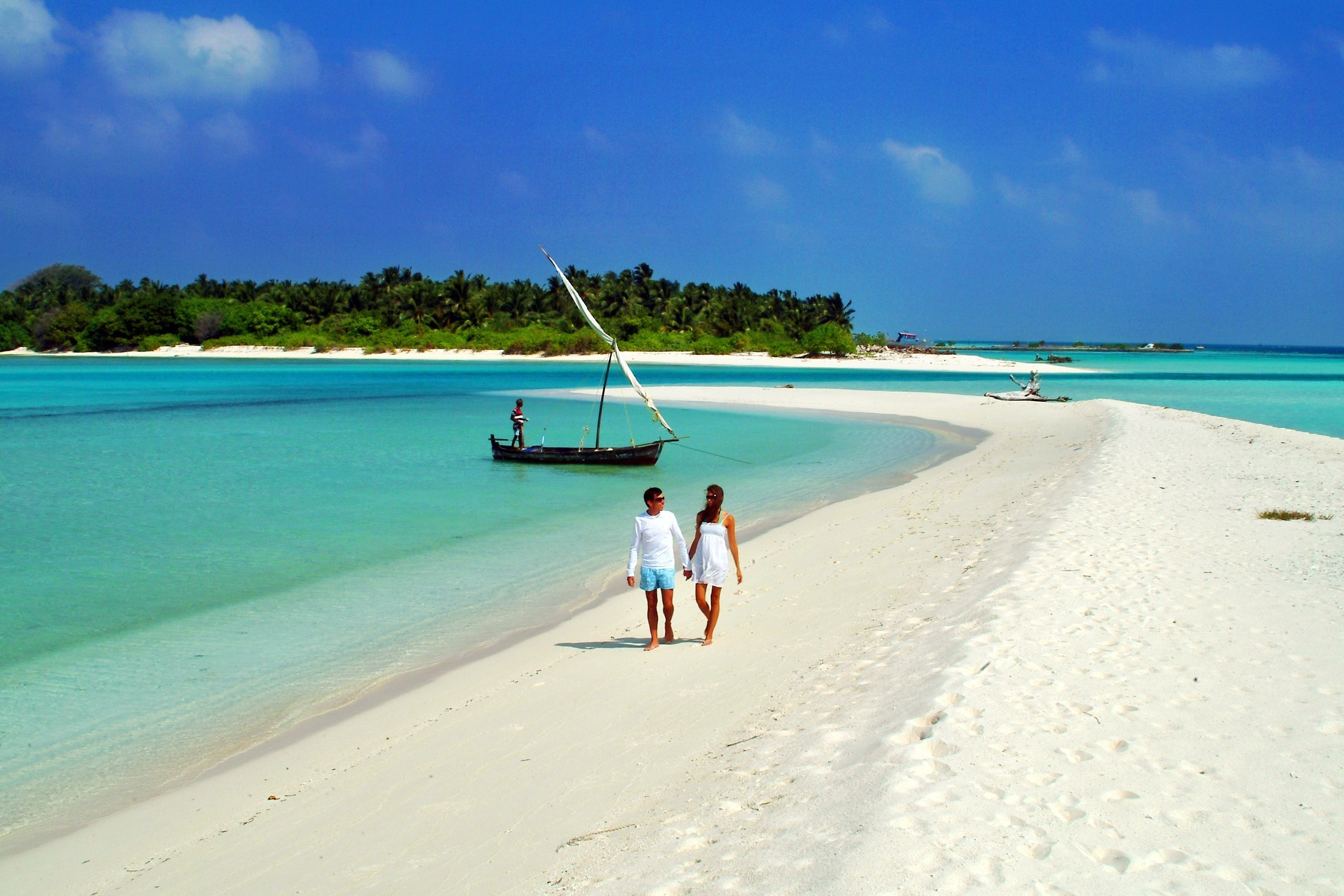 A beach with clear turquoise water, a boat, and a couple walking on the sand.